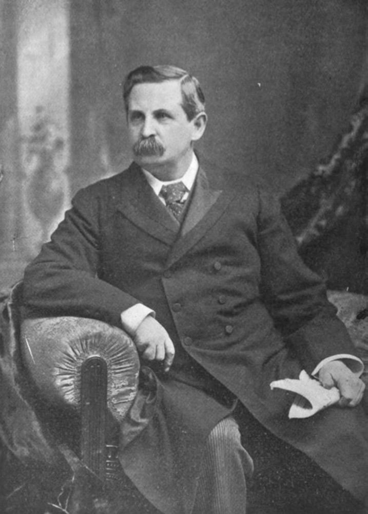 Black-and-white 3/4-photographic image of a moustached man in a long suit-coat and tie. He is seated in a chair, gazing off past the camera’s left. His left hand holds a newspaper and his right arm rests on the chair arm. The pose, gaze, and a background panel possibly evoking a classical motif suggests this is a studio portrait.