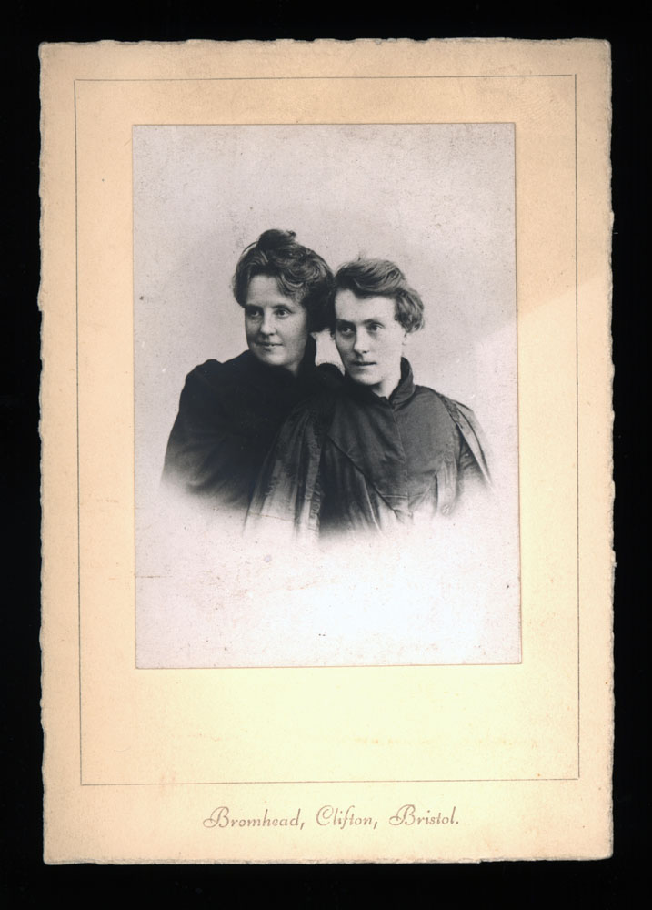 This photograph of Katherine Bradley and Edith Cooper is taken from the chest up fading out around the mid-chest area. Both women are positioned with their bodies angled slightly to the right, and are looking towards the left side of the photograph. Katherine is in the front, wearing a high-necked dress with a cape or jacket hanging on her shoulders. Her hair is short, covering the tops of her ears. It’s parted on the right side of her head, and is swept back. Edith is also wearing a high-necked dress, details are obscured in shadow. Her hair is up in a bun. Both women are slightly smiling. The photo is on a plain card, which reads “Bromhead, Clifton, Bristol” in cursive typeface at the bottom.
