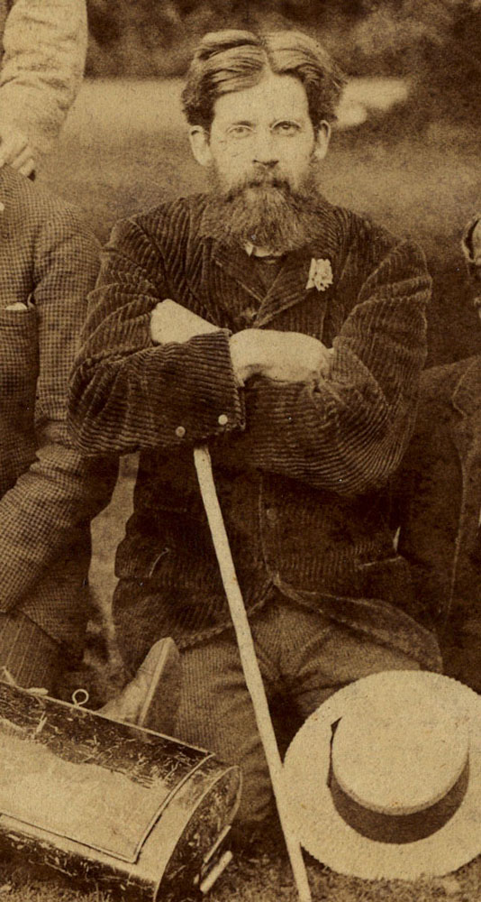 Sepia-tone image is of Patrick Geddes on a field study, kneeling on the ground. His arms are crossed and his gaze is directly on the viewer. He is wearing a dark corduroy suit jacket and lighter pants. He is holding a white stick in one hand; possibly a walking stick. A circular-brimmed light hair lies on the ground, covering his left knee and thigh.