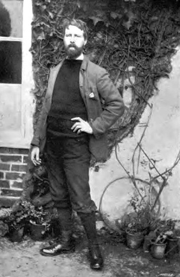 This photograph of Thomas Cooper Gotch is full-length and shows him standing outside of a building, facing the camera with his left hand on his hip, right hand hanging loosely. His left foot points forwards, and right foot points left. He is wearing a sweater, trousers, and jacket; the left side of his jacket rests behind his left arm. A light handkerchief is sticking out of the left breast pocket. He has a full beard, and his hair is cut short, about 2 inches long. One piece of hair has been swept onto his forehead. The photograph was taken beside a building, in the background the brick wall, a large ivy vine and window are visible. Potted plants are on the ground behind him, on either side of him.