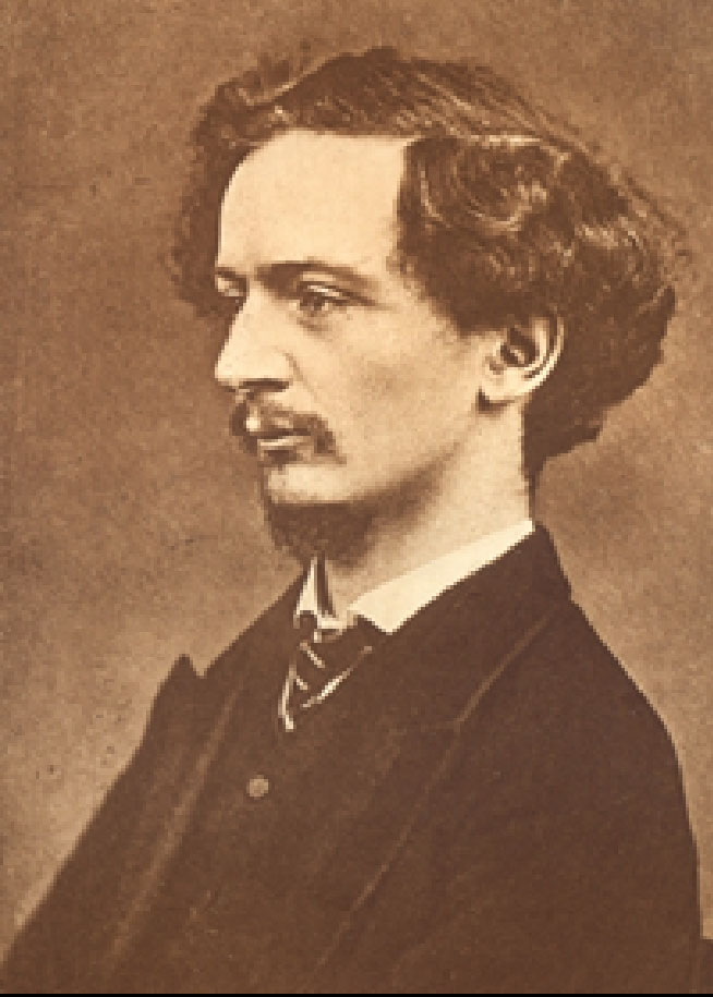 Image is a photograph of Algernon Charles Swinburne shown from the shoulders up. He is wearing a dark suit jacket, a white shirt and a striped tie. He is shown in profile looking to the left. His hair is wavy and parted on the right side. He has a moustache. The background is dark and undefined. The image is vertically displayed.