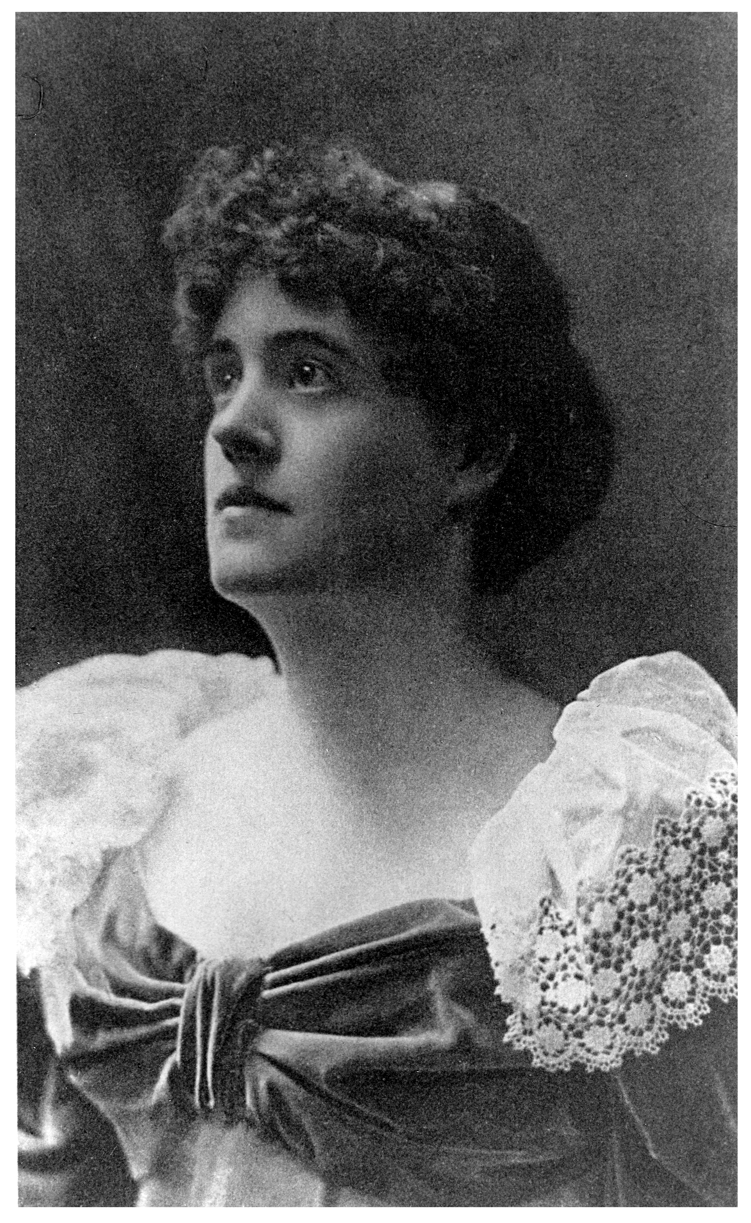 Image is a photograph of Graham Tomson [Rosamund Marriott Watson] shown from the chest up. She is in ¾ face, looking towards the upper left corner of the image. She has curly hair that is worn short. She is wearing a dress with a low neckline; the fabric gathers in a bow in the middle of her chest. The dress has long sleeves accentuated with white lace shoulders. The background is open and dark-coloured. The image is vertically displayed.