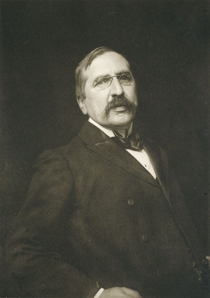 Gleeson White is shown facing forwards from the waist up. He is looking straight ahead. He is wearing a dark double breasted suit, a white collared shirt and a black satin bow tie. His right hand appears to be resting at his side with his elbow slightly bent. His hair is combed to the right side and he is sporting a handlebar moustache and metal rimmed ‘pince nez’ spectacles.
