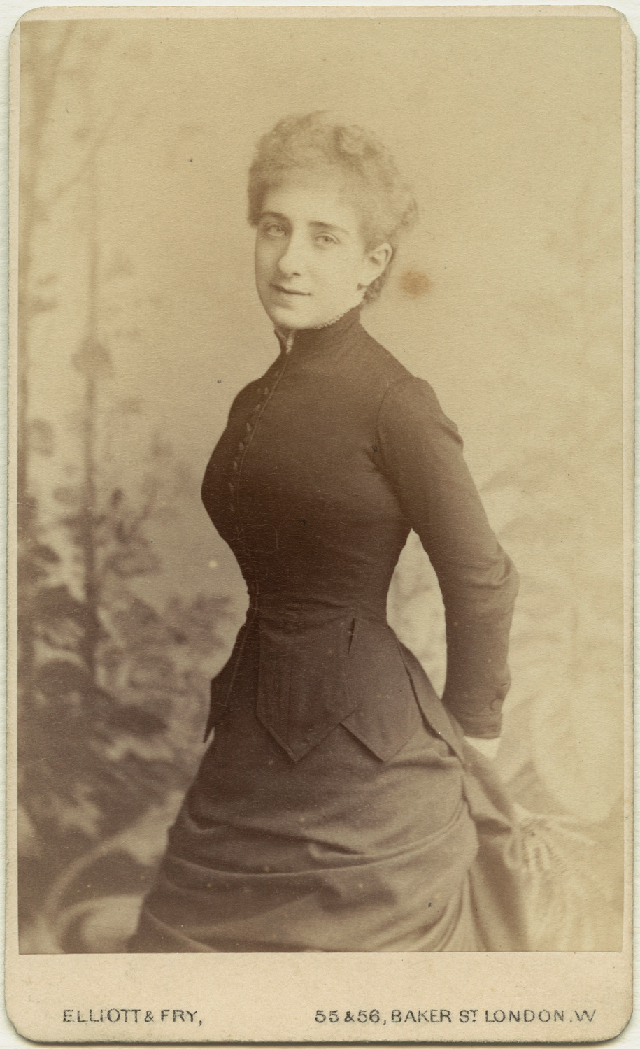 Ada Leverson is shown standing (or possibly leaning) with hands clasped behind her in a front facing pose, angled slightly to the right. Though her body is angled, she is gazing straightforward. Her eyes appear to be lightly coloured, and she is smiling softly. Her hair is very short, curly, and falls slightly onto her forehead. Leverson is wearing a dark button-up dress. The dress has a collar that extends the length of her neck, decorative pointed fabric that hangs off her waist section, and a skirt that extends outward at her waist and bunches up behind her. In the background, behind Leverson, there appears to be a garden with upward growing vines. The image is shaded in brown sepia tones, and has a beige rounded-rectangular border. The text “ELLIOTT & FRY 55&56, BAKER ST LONDON, W” is written in capitalized letters within the bottom border of the photograph.