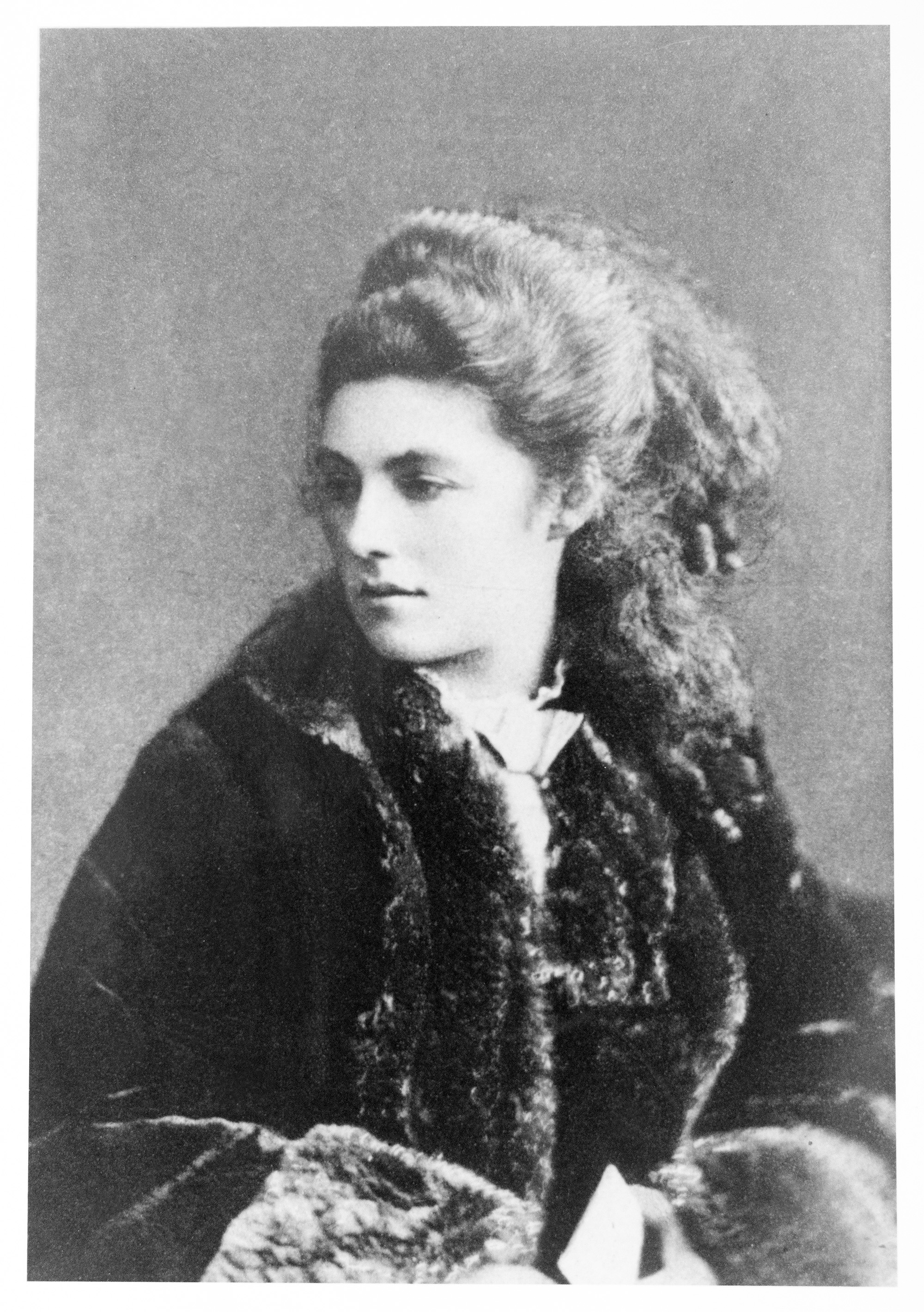 This photograph of Mathilde Blind shows her seated, and is taken from the waist up. Her head is turned slightly to the right, her body angled the left. She is wearing a large fur coat, and her sleeved arms are visible at the bottom of the photograph. She is holding a print object. Her hair is half pinned up, left ear visible, and the ends are curled into small ringlets.