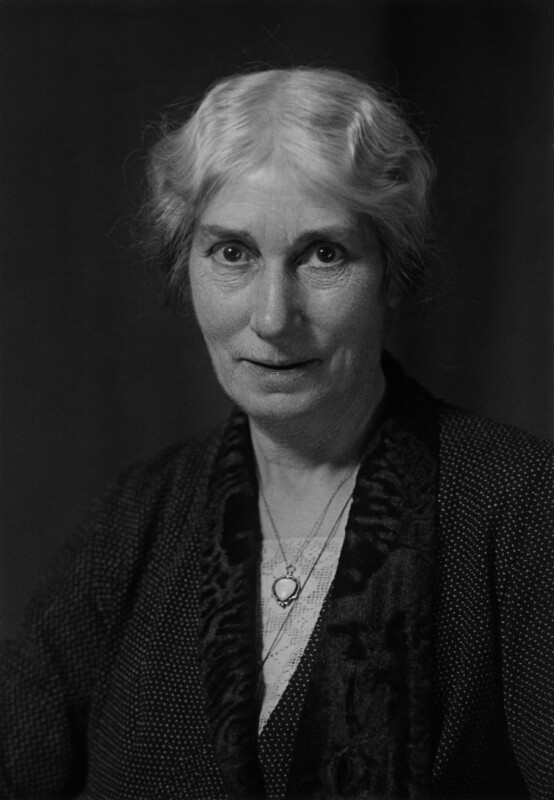 The photograph is in black and white. Evelyn Sharp is shown in a                            head-and-shoulders pose with her body slightly angled towards the left                            of the image. Her gaze is straight on the camera, directed at the                            viewer, and she is slightly smiling. Her hair is grey or white, has a                            softly waved texture, and is tied back, partially covering her ears. She                            is wearing a dark dress with a tiny polka dot print. There is a                            triangular lace cutout at the collar of her shirt, and she is wearing a                            scarf with a velvety texture. She is wearing two necklaces, one has a                            pendant with an upside-down teardrop shaped stone inlaid. The other                            necklace is partially obscured by her scarf, no pendant is                            visible.