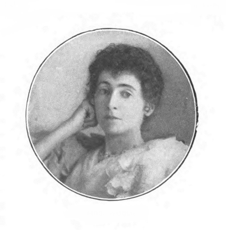 Margaret Louisa Woods is shown in a head-and-shoulders pose. Her body is facing right, but her head and gaze are directed straightforward. Her right arm is bent, and her right hand is making a relaxed fist positioned up against her cheek. Her facial features are illuminated by a light source coming in from the right side of the image, casting a minimal shadow on the left side of her face. She is not smiling, but her eyebrows are slightly raised. Woods’ hair is dark, curly, and cut short to frame her face. She is wearing ball-stud earrings and a beaded necklace. She is wearing a ¾ sleeve chiffon dress that is lightly coloured. The photograph is coloured black and white, and positioned within a circular lined frame.