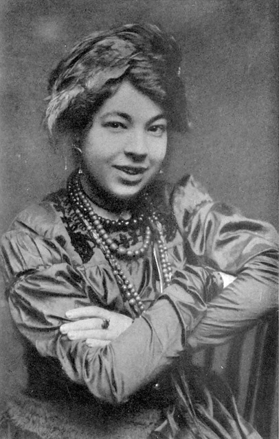 Pamela Colman Smith is shown seated, from the waist up. One arm rests on the back of a wooden chair, and both are crossed in front. Her mouth is open in a smile, and her hair is in an updo with an ornament pinned to the side. She is wearing 4 beaded necklaces of varying lengths and colours, and a ring on her left middle finger. Her dress is embroidered in the collar area, and has puffed sleeves that tighten from the elbows to the wrists.