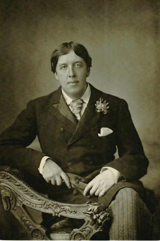 A young Oscar Wilde sits sideways facing the camera on an ornate bench. His left leg is crossed over his right. He is leaning his right elbow on the back of the bench and holding a pair of gloves between both hands. He is wearing pin-striped pants and a long jacket buttoned to the top with a tie visible underneath. There is a white kerchief tucked into his left breast pocket and a flower pinned to his left lapel.